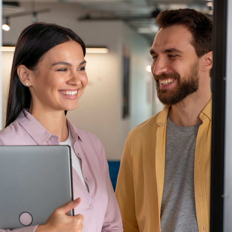Two employees in casual outfits smiling and looking at a laptop